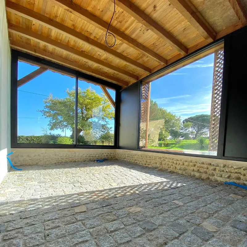 Coulissant en angle à cadres noirs posé dans une dépendance au plafond en bois, ouvert sur le jardin et un grand arbre