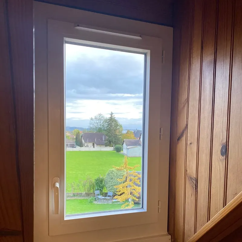 Fenêtre PVC blanche à un vantail posée dans une pièce lambrissée, vue sur un pré