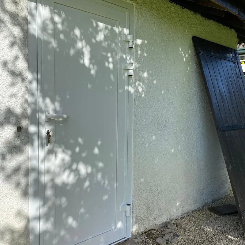Après : Porte pleine claire posée sur le mur blanc d’un abri, l’ancienne porte en bois appuyée à côté